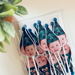 Puede incluir: Un conjunto de adornos para pasteles con la foto de un ni&ntilde;o peque&ntilde;o que lleva un gorro de fiesta a cuadros verdes y azules con el n&uacute;mero 1 impreso. Cada adorno est&aacute; unido a un palito blanco e incluye un peque&ntilde;o pomp&oacute;n azul.
