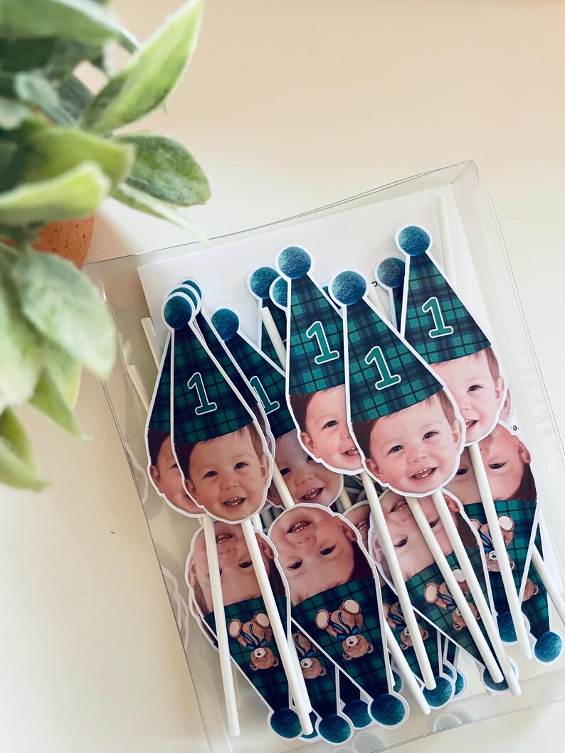 Puede incluir: Un conjunto de adornos para pasteles con la foto de un ni&ntilde;o peque&ntilde;o que lleva un gorro de fiesta a cuadros verdes y azules con el n&uacute;mero 1 impreso. Cada adorno est&aacute; unido a un palito blanco e incluye un peque&ntilde;o pomp&oacute;n azul.