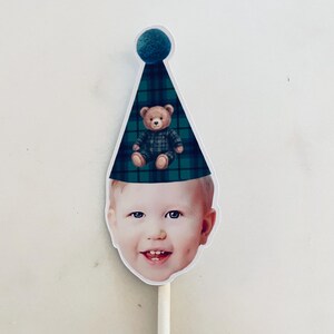 Puede incluir: Un recorte de la cara de un ni&ntilde;o con un gorro de fiesta de cuadros verdes y azules con un pomp&oacute;n azul verdoso en la parte superior. Un peque&ntilde;o oso de peluche marr&oacute;n con un conjunto de cuadros a juego est&aacute; impreso en el gorro. Unido a un palo blanco.