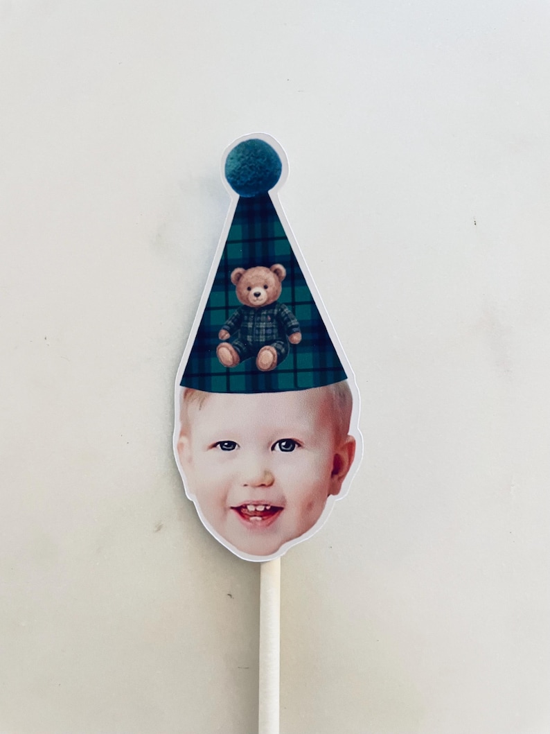 Puede incluir: Un recorte de la cara de un ni&ntilde;o con un gorro de fiesta de cuadros verdes y azules con un pomp&oacute;n azul verdoso en la parte superior. Un peque&ntilde;o oso de peluche marr&oacute;n con un conjunto de cuadros a juego est&aacute; impreso en el gorro. Unido a un palo blanco.