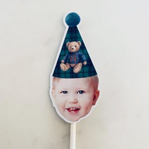 Puede incluir: Un recorte del rostro de un ni&ntilde;o sonriente que lleva un gorro de fiesta a cuadros verde oscuro y azul con un pomp&oacute;n azul. Un peque&ntilde;o oso de peluche marr&oacute;n con un su&eacute;ter azul estampado est&aacute; impreso en el frente del gorro. El recorte est&aacute; unido a un palo.