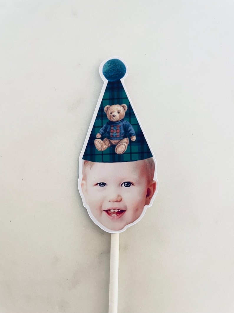 Puede incluir: Un recorte del rostro de un ni&ntilde;o sonriente que lleva un gorro de fiesta a cuadros verde oscuro y azul con un pomp&oacute;n azul. Un peque&ntilde;o oso de peluche marr&oacute;n con un su&eacute;ter azul estampado est&aacute; impreso en el frente del gorro. El recorte est&aacute; unido a un palo.