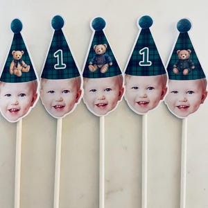 Puede incluir: Cinco adornos para pastel con el rostro de un ni&ntilde;o que lleva un gorro de fiesta a cuadros verdes y azules con un pomp&oacute;n. Algunos gorros muestran el n&uacute;mero uno, mientras que otros tienen una ilustraci&oacute;n de un osito. Cada adorno tiene un palito.