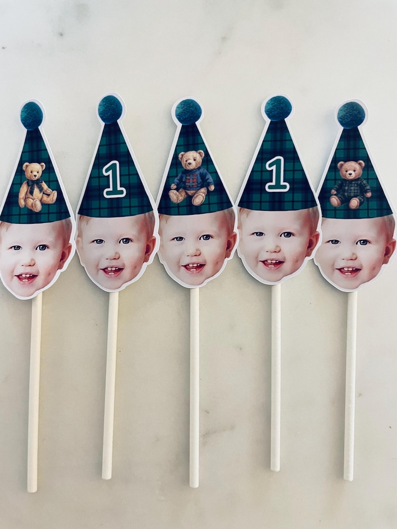 Puede incluir: Cinco adornos para pastel con el rostro de un ni&ntilde;o que lleva un gorro de fiesta a cuadros verdes y azules con un pomp&oacute;n. Algunos gorros muestran el n&uacute;mero uno, mientras que otros tienen una ilustraci&oacute;n de un osito. Cada adorno tiene un palito.