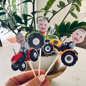 May include: Three cupcake toppers featuring a child's face on a tractor. The tractors are red, green, and yellow. The toppers are on white sticks.