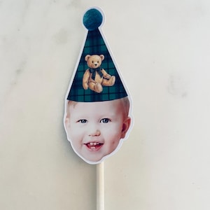 Puede incluir: Un adorno para pastel con el recorte de la cara de un ni&ntilde;o sonriente que lleva un gorro de fiesta a cuadros verdes y azules con un pomp&oacute;n azul oscuro y una ilustraci&oacute;n de un osito de peluche marr&oacute;n, sujeto a un palo blanco.