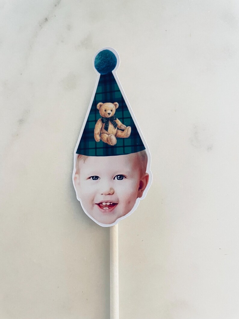 Puede incluir: Un adorno para pastel con el recorte de la cara de un ni&ntilde;o sonriente que lleva un gorro de fiesta a cuadros verdes y azules con un pomp&oacute;n azul oscuro y una ilustraci&oacute;n de un osito de peluche marr&oacute;n, sujeto a un palo blanco.