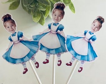 May include: Three Alice in Wonderland-themed cupcake toppers. Each topper features a watercolor illustration of a girl in a blue dress, white apron, and red shoes. The toppers are attached to white lollipop sticks, perfect for a themed party.