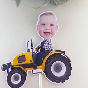 May include: A yellow toy tractor with a black and white tyre design. A baby's face is on the tractor, wearing a blue and white plaid shirt and blue overalls. The tractor is on a white stick.