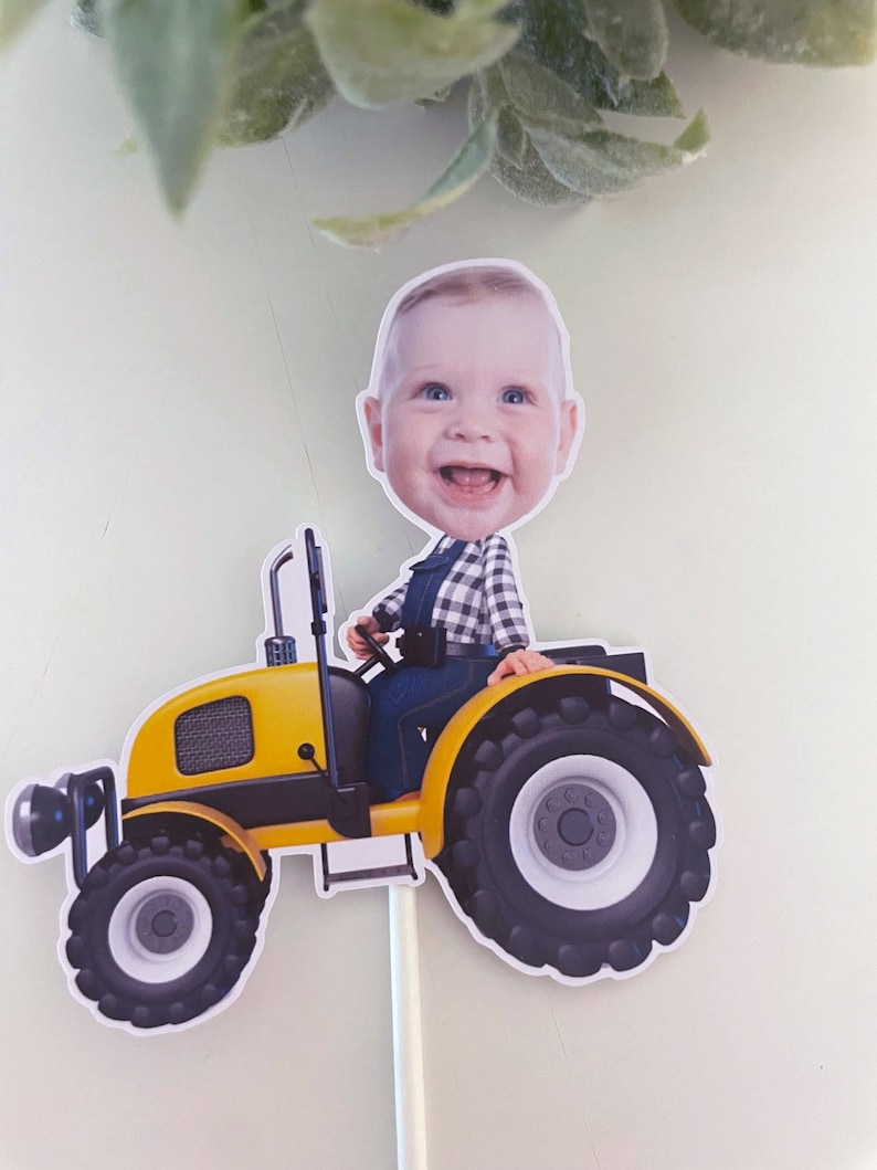 May include: A yellow toy tractor with a black and white tyre design. A baby's face is on the tractor, wearing a blue and white plaid shirt and blue overalls. The tractor is on a white stick.