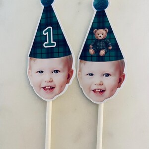 Puede incluir: Dos adornos personalizados para tarta con la cara recortada de un ni&ntilde;o sonriente sobre un palo de madera. Cada uno lleva un gorro de fiesta de cuadros verdes y azules con un pomp&oacute;n azul. Un gorro muestra el n&uacute;mero 1 y el otro un osito.