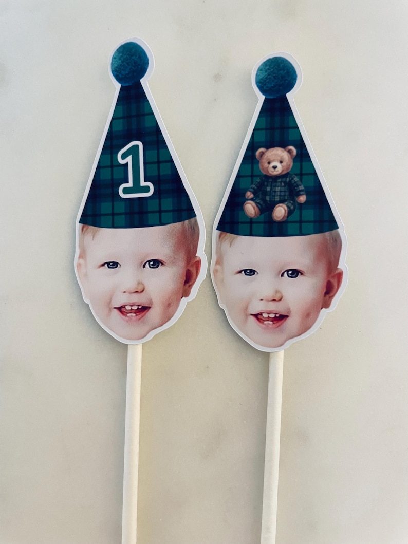 Puede incluir: Dos adornos personalizados para tarta con la cara recortada de un ni&ntilde;o sonriente sobre un palo de madera. Cada uno lleva un gorro de fiesta de cuadros verdes y azules con un pomp&oacute;n azul. Un gorro muestra el n&uacute;mero 1 y el otro un osito.