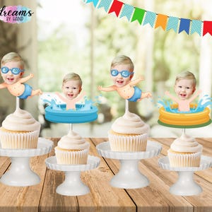 May include: Four cupcakes on white pedestals, each topped with a paper cutout. Two feature a baby wearing blue goggles and swimming, while two show a baby in a small inflatable pool. The text "dreams BY HAND" appears in the upper left corner.