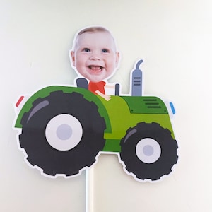 May include: A green toy tractor with a baby's face on top. The tractor has a black steering wheel, black wheels, and a grey exhaust pipe.