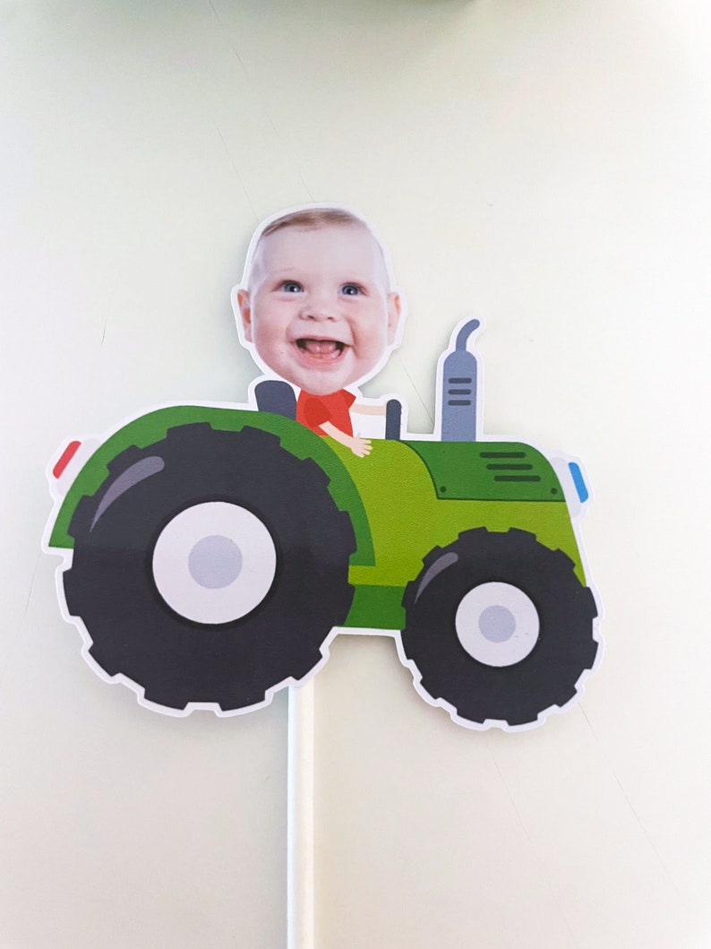 May include: A green toy tractor with a baby's face on top. The tractor has a black steering wheel, black wheels, and a grey exhaust pipe.