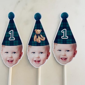 Puede incluir: Tres adornos personalizados para pasteles con el rostro de un ni&ntilde;o que lleva un gorro de fiesta a cuadros verdes y azules con un pomp&oacute;n verde azulado. Dos gorros muestran el n&uacute;mero 1, mientras que el central tiene un osito de peluche.