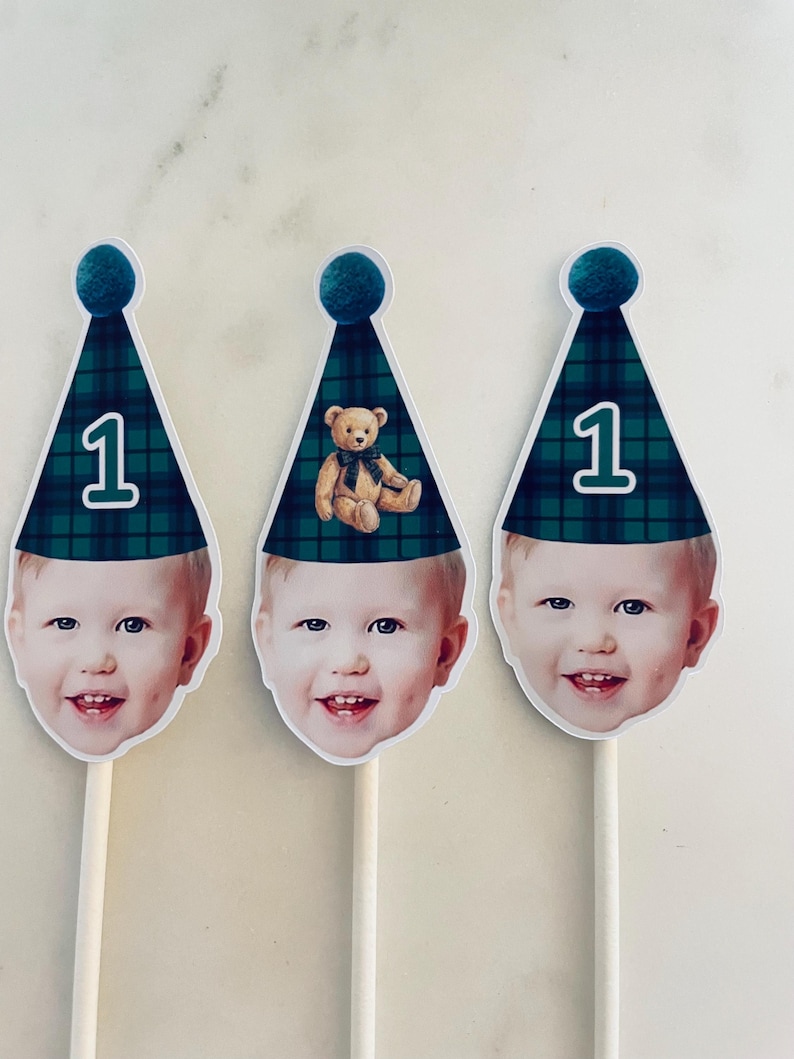 Puede incluir: Tres adornos personalizados para pasteles con el rostro de un ni&ntilde;o que lleva un gorro de fiesta a cuadros verdes y azules con un pomp&oacute;n verde azulado. Dos gorros muestran el n&uacute;mero 1, mientras que el central tiene un osito de peluche.