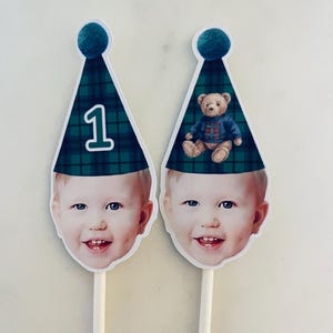 Puede incluir: Dos adornos para tarta con la cara de un ni&ntilde;o recortada sobre un palito. Cada adorno tiene un gorro de fiesta c&oacute;nico de cuadros verdes y azules con un pomp&oacute;n azul. Uno muestra el n&uacute;mero 1 y el otro un oso de peluche.