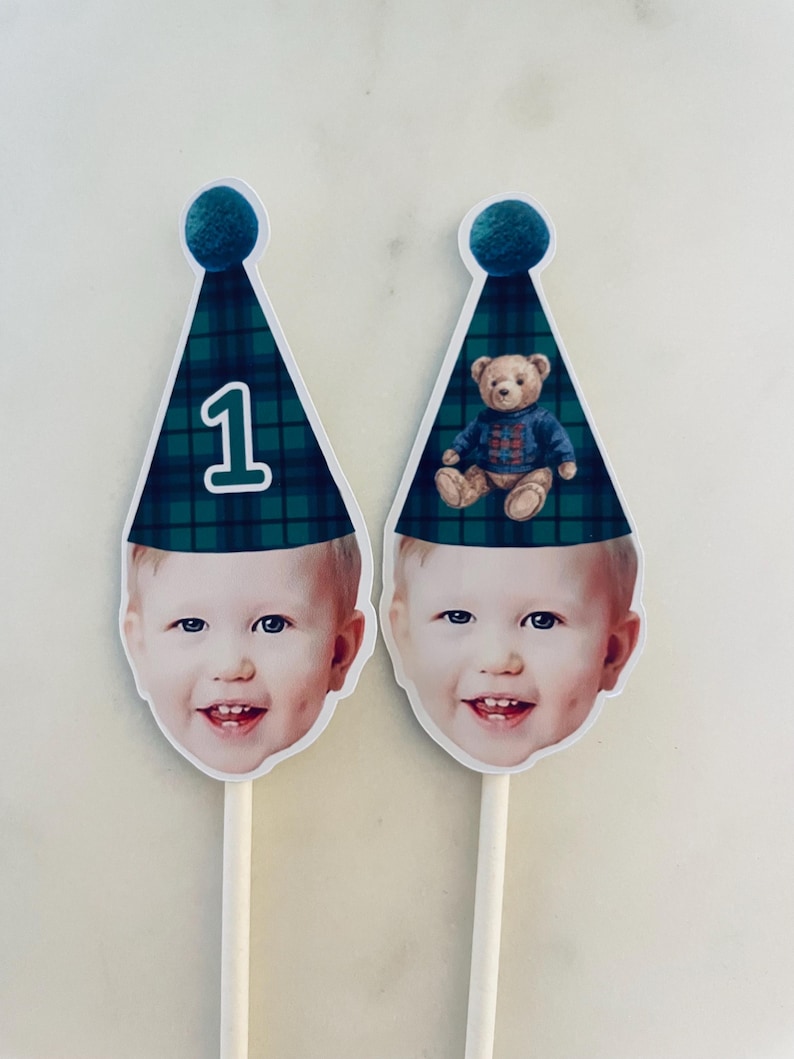 Puede incluir: Dos adornos para tarta con la cara de un ni&ntilde;o recortada sobre un palito. Cada adorno tiene un gorro de fiesta c&oacute;nico de cuadros verdes y azules con un pomp&oacute;n azul. Uno muestra el n&uacute;mero 1 y el otro un oso de peluche.