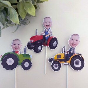 May include: Three cupcake toppers featuring a child's face on a tractor. The tractors are red, green, and yellow. The toppers are perfect for a farm-themed birthday party.