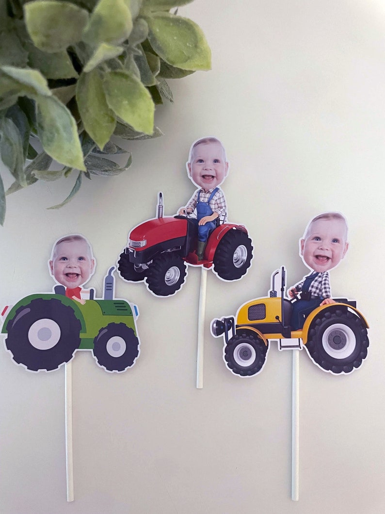 May include: Three cupcake toppers featuring a child's face on a tractor. The tractors are red, green, and yellow. The toppers are perfect for a farm-themed birthday party.