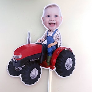 May include: A red toy tractor with a smiling baby's head on top, wearing blue overalls and a plaid shirt. The tractor has large black wheels and a white stick attached to the bottom.