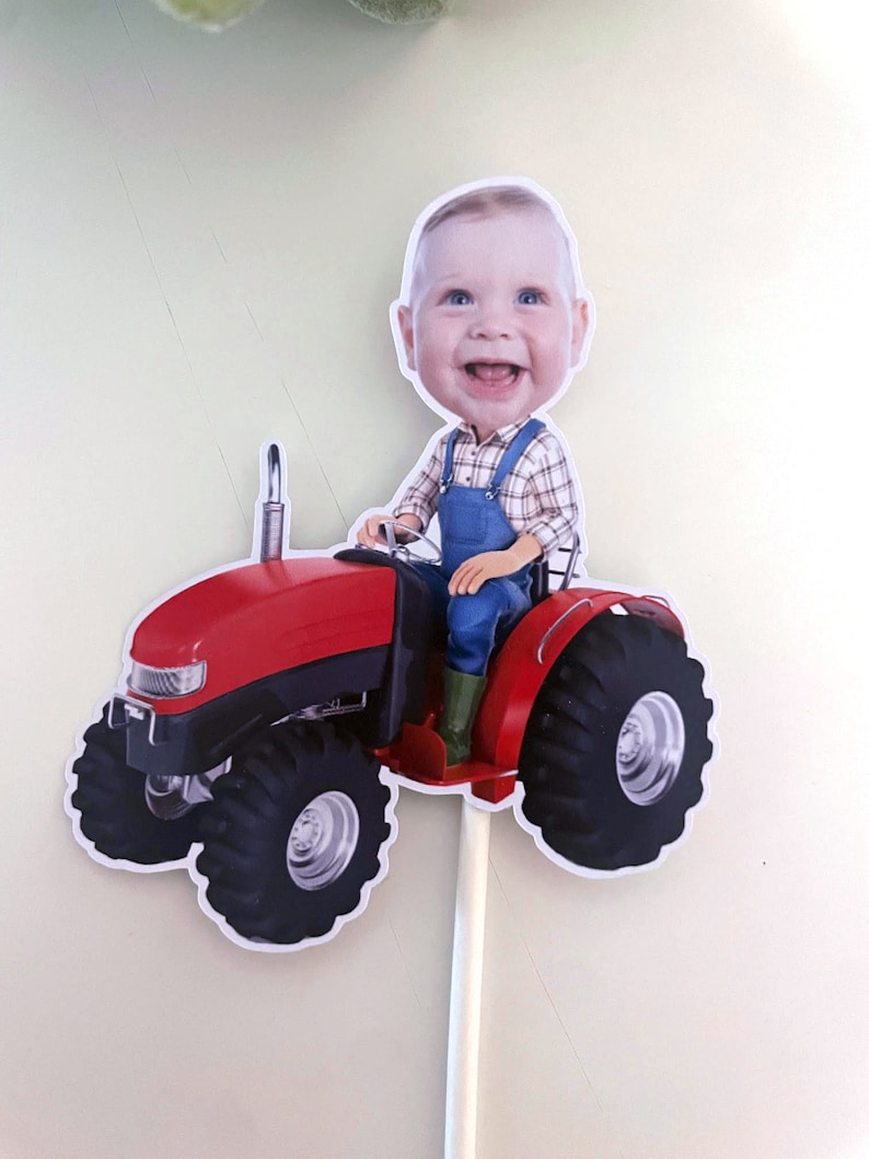 May include: A red toy tractor with a smiling baby's head on top, wearing blue overalls and a plaid shirt. The tractor has large black wheels and a white stick attached to the bottom.