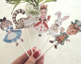 May include: A set of five decorative paper toppers on white sticks featuring illustrated characters from Alice in Wonderland, including a cat, a rabbit, and three figures with human faces integrated into the designs, showcasing colorful outfits.