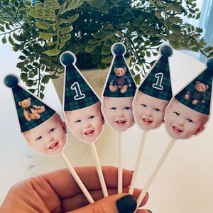 Puede incluir: Cinco adornos para cupcakes con la cara de un ni&ntilde;o que lleva un gorro de fiesta de cuadros verdes y azules. Cada gorro tiene un pomp&oacute;n azul y muestra el n&uacute;mero 1 o un peque&ntilde;o oso de peluche. Los adornos est&aacute;n sujetos a palitos blancos.