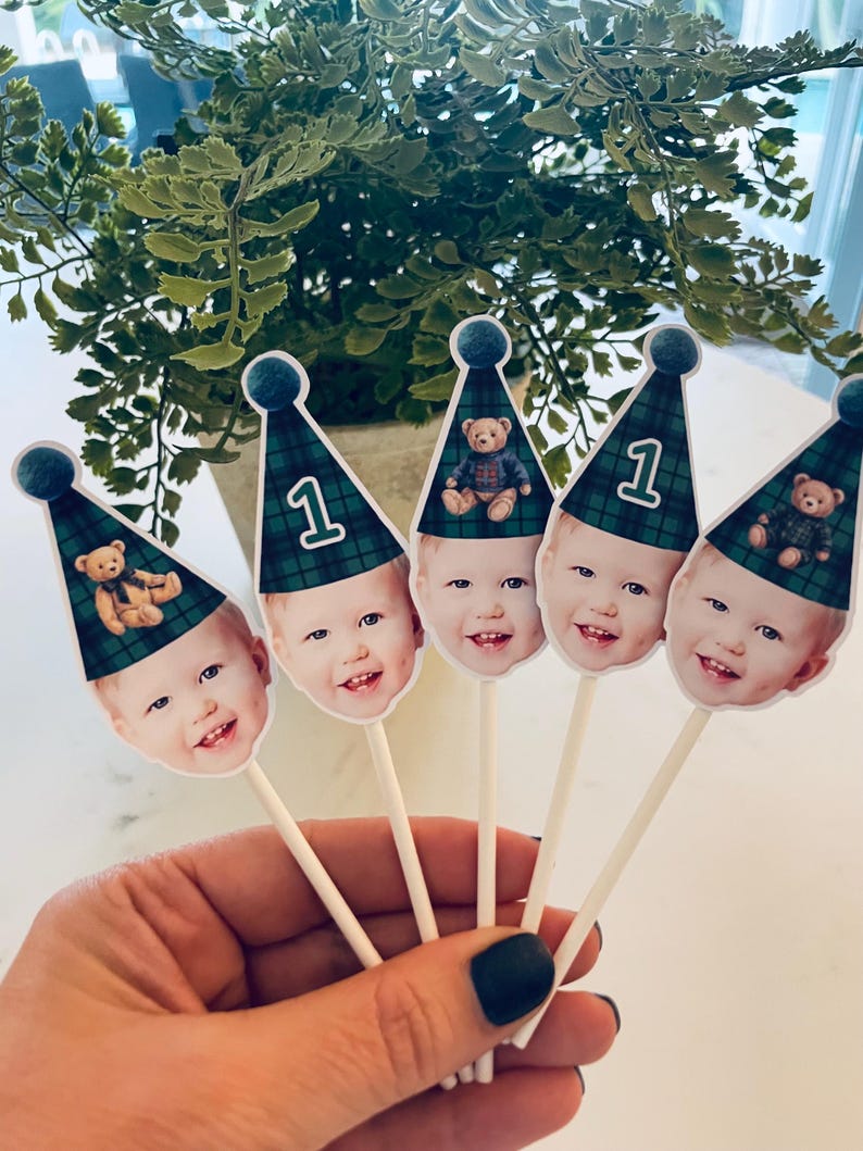 Puede incluir: Cinco adornos para cupcakes con la cara de un ni&ntilde;o que lleva un gorro de fiesta de cuadros verdes y azules. Cada gorro tiene un pomp&oacute;n azul y muestra el n&uacute;mero 1 o un peque&ntilde;o oso de peluche. Los adornos est&aacute;n sujetos a palitos blancos.