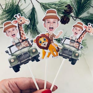 Puede incluir: Tres adornos para cupcakes con temática de safari con imágenes de dibujos animados de un león, una jirafa y una persona con un sombrero de safari. Cada adorno está montado en un palo blanco y presenta un jeep verde.