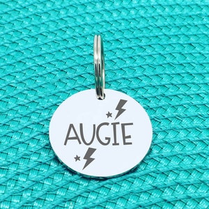 May include: A round, silver pet tag with the name "AUGIE" in a bold font. The tag features a lightning bolt and star design. A silver split ring is attached at the top for easy attachment to a pet's collar. The tag is set against a turquoise background.