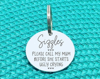 May include: A round silver-toned metal pet ID tag with a split ring. The tag is engraved with the name Sizzles, followed by the text: PLEASE CALL MY MUM BEFORE SHE STARTS UGLY CRYING, accented with small heart symbols above and below the text.