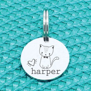 May include: A round, silver pet tag with a cartoon cat illustration, a heart, and the name "harper" in a simple font. The tag has a silver ring for attachment. The background is a textured turquoise surface.