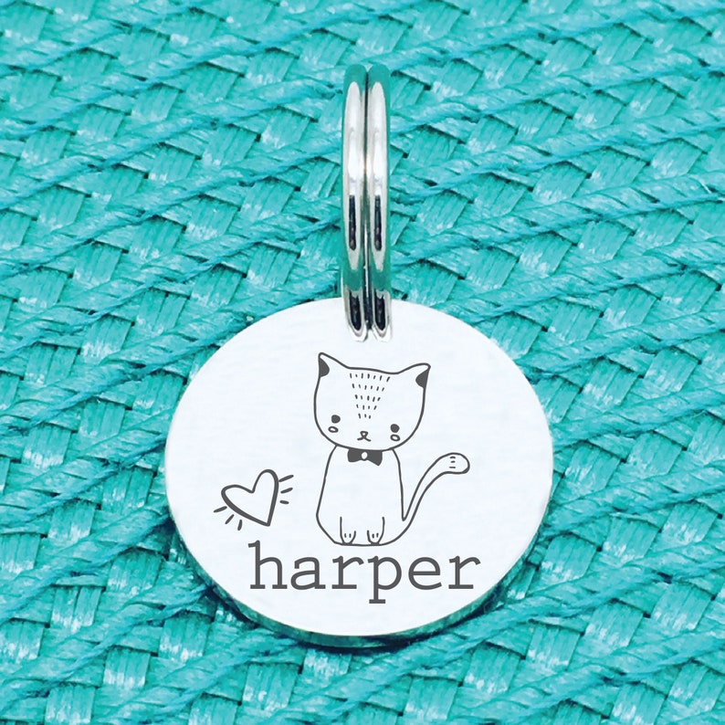 May include: A round, silver pet tag with a cartoon cat illustration, a heart, and the name "harper" in a simple font. The tag has a silver ring for attachment. The background is a textured turquoise surface.