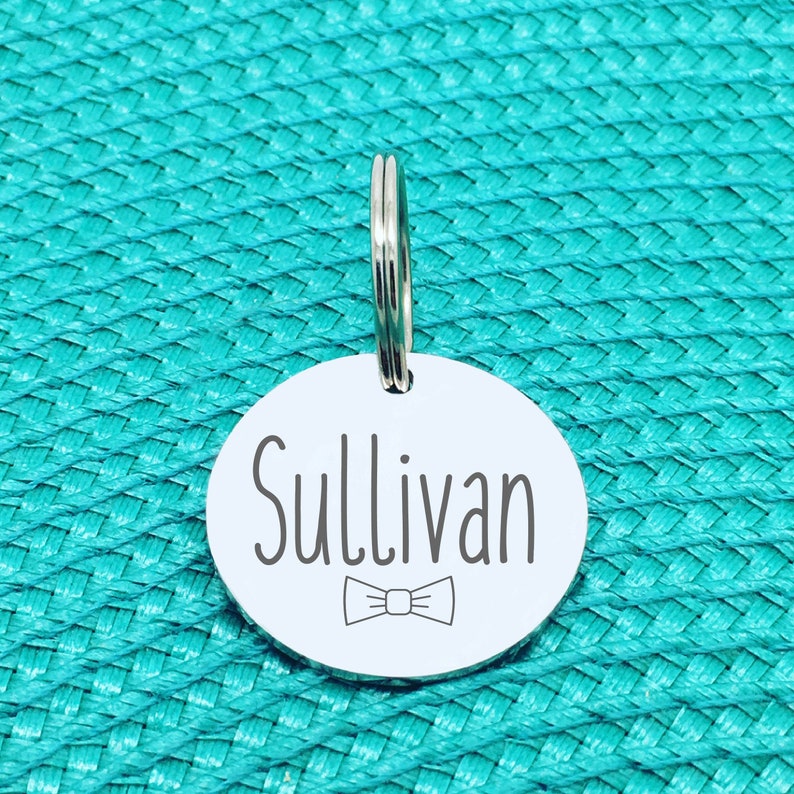 May include: A round, silver pet tag with the name "Sullivan" in a dark gray font, along with a bow tie graphic. The tag is attached to a silver split ring. The background is a textured turquoise surface.