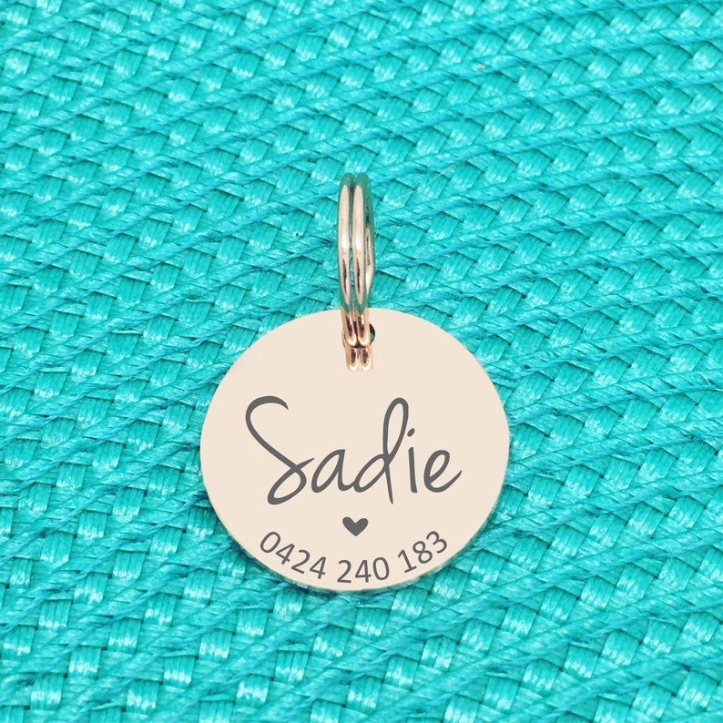 K&ouml;nnte beinhalten: Ein ros&eacute;goldfarbener Hundeanh&auml;nger mit dem Namen "Sadie" in Schreibschrift, einem kleinen Herz und einer Telefonnummer. Der Anh&auml;nger ist rund und hat einen Ring zur Befestigung. Auf t&uuml;rkisfarbenem Hintergrund.