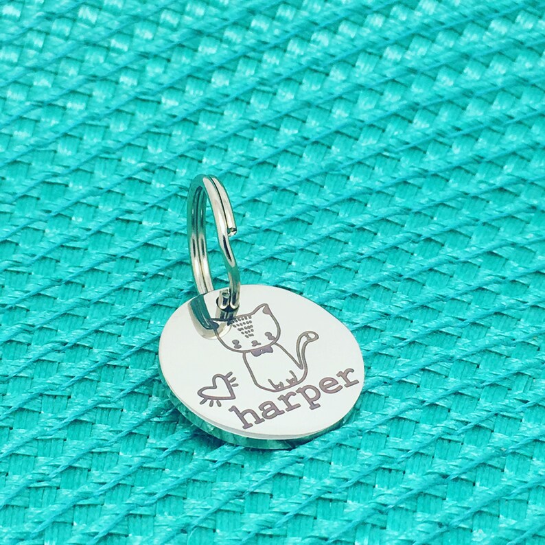 May include: A silver-colored, circular pet tag with a cat illustration, a heart, and the name "harper" engraved in a dark color. The tag is attached to a silver key ring. The background is a textured teal surface.