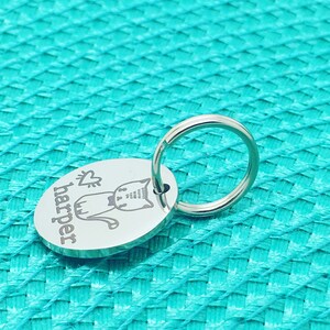 May include: A silver-colored, oval-shaped keychain with a cat illustration and the name "Harper" engraved on it. The keychain has a small heart symbol above the cat. The background is a textured, turquoise surface.
