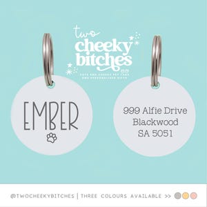 May include: Two white pet tags with silver rings. One tag has the name "EMBER" and a paw print. The other tag has an address. The text "two cheeky bitches" is at the top.
