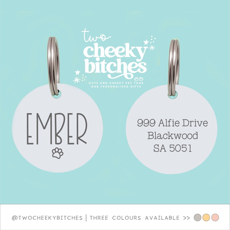 May include: Two white pet tags with silver rings. One tag has the name "EMBER" and a paw print. The other tag has an address. The text "two cheeky bitches" is at the top.