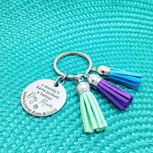 May include: A silver keychain with a round charm engraved with "I couldn't have picked a better Dog Mum" and a paw print. It has three colorful tassels in blue, purple, and green. The keychain is on a turquoise textured surface.