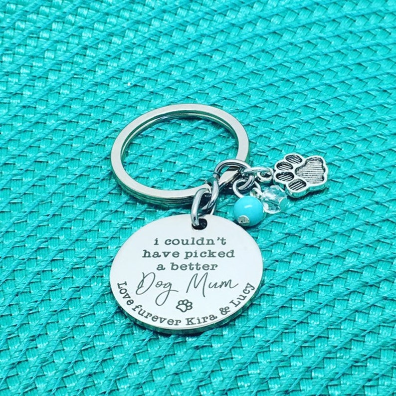 May include: A silver-colored keychain with a round pendant that reads "I couldn't have picked a better Dog Mum." The keychain has a paw print charm, a blue bead, and a clear crystal charm. The words "Love furever Kira & Lucy" are also on the pendant.