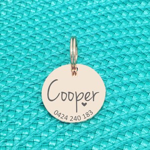 K&ouml;nnte beinhalten: Ein ros&eacute;goldener, runder Haustieranh&auml;nger mit dem Namen "Cooper" in handschriftlicher Schrift, einem kleinen Herz und einer Telefonnummer. Der Anh&auml;nger ist an einem silbernen Ring befestigt. Der Hintergrund ist eine strukturierte t&uuml;rkisfarbene Oberfl&auml;che.