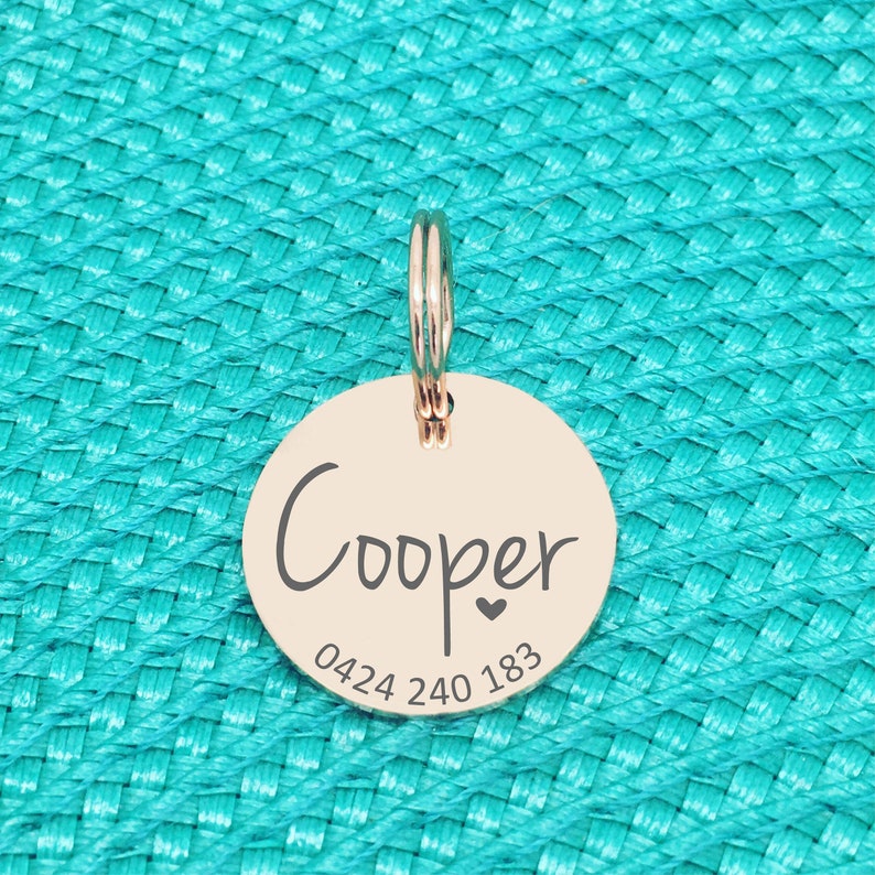 K&ouml;nnte beinhalten: Ein ros&eacute;goldener, runder Haustieranh&auml;nger mit dem Namen "Cooper" in handschriftlicher Schrift, einem kleinen Herz und einer Telefonnummer. Der Anh&auml;nger ist an einem silbernen Ring befestigt. Der Hintergrund ist eine strukturierte t&uuml;rkisfarbene Oberfl&auml;che.