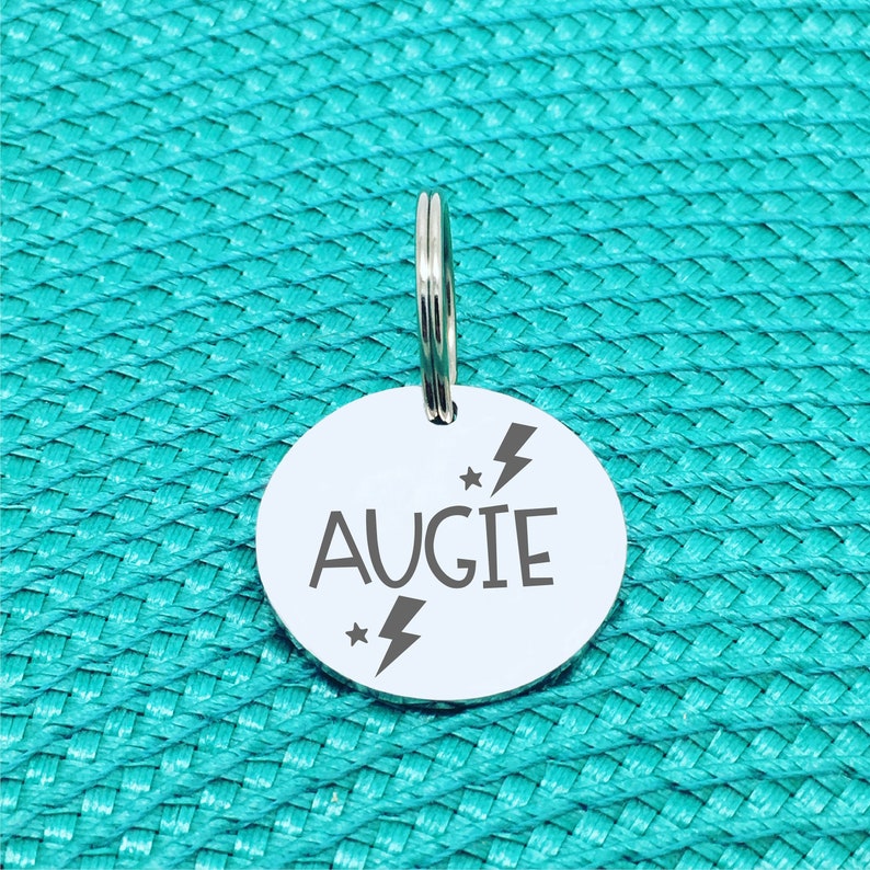 May include: A round, silver pet tag with the name "AUGIE" in a bold font. The tag features a lightning bolt and star design. A silver split ring is attached at the top for easy attachment to a pet's collar. The tag is set against a turquoise background.