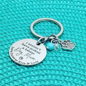 May include: A silver-colored keychain with a round pendant engraved with the words "I couldn't have picked a better Dog Mum" and the names Kira & Lucy. It includes a paw print charm, a turquoise bead, and a clear crystal charm.