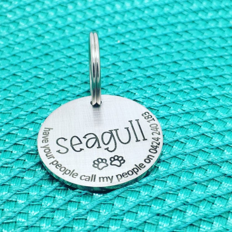May include: A round, silver pet tag with the name "seagull" in a bold font. The tag includes paw prints and the phrase "have your people call my people on 0424 240 183". A silver ring is attached for easy attachment to a collar.