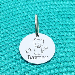 May include: A round, silver pet tag with a cat illustration and the name "Baxter" engraved in a simple font. The cat has a bow tie and a heart next to it. A silver ring is attached at the top for easy attachment to a collar.