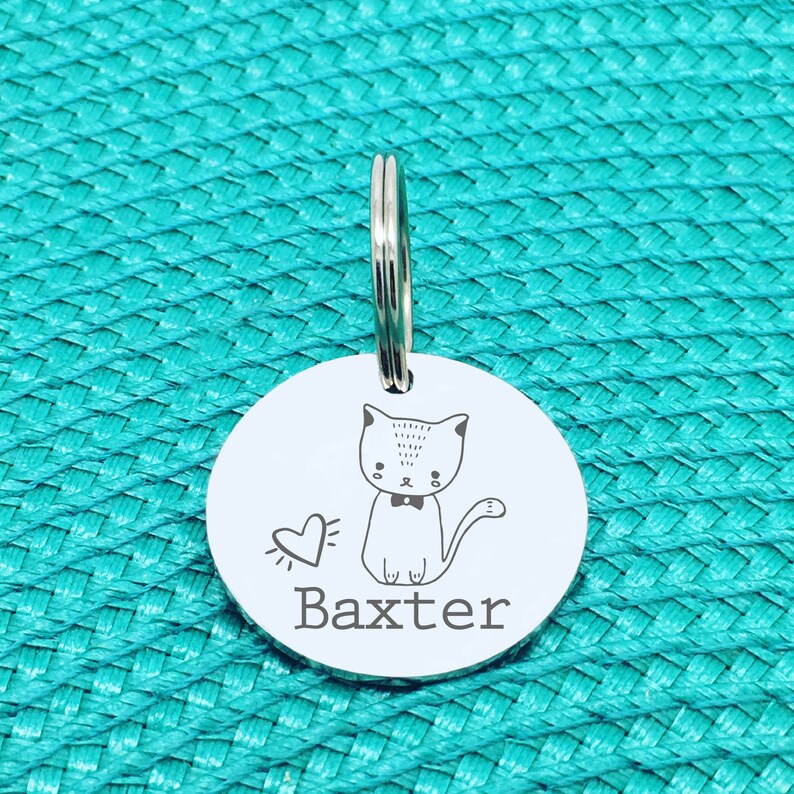 May include: A round, silver pet tag with a cat illustration and the name "Baxter" engraved in a simple font. The cat has a bow tie and a heart next to it. A silver ring is attached at the top for easy attachment to a collar.